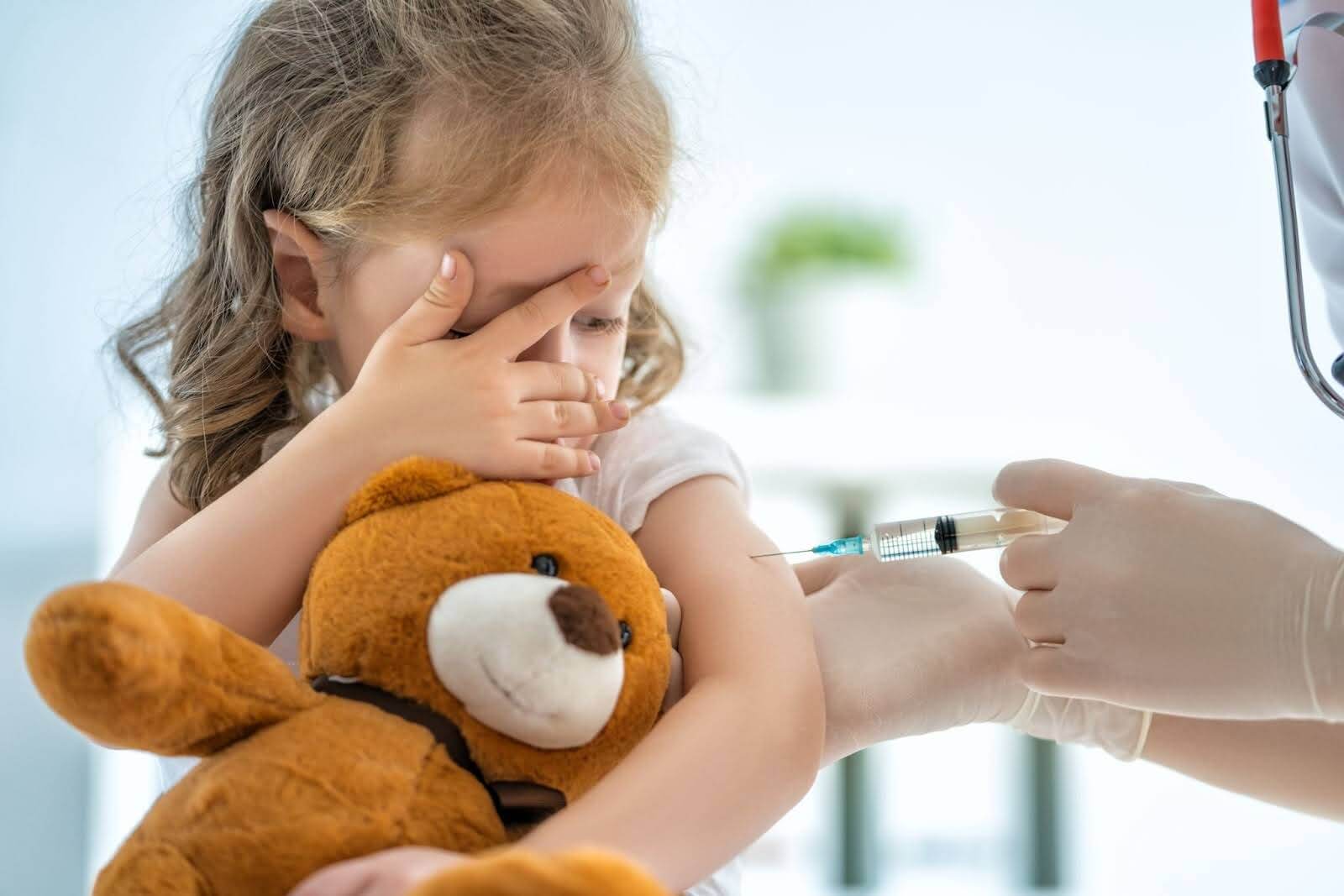 A little girl holding a teddybear while being vaccinated