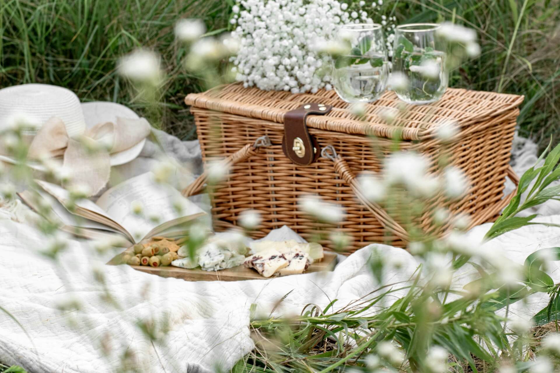 A picnic basket, food, and two glasses with water on a blanket on the grass